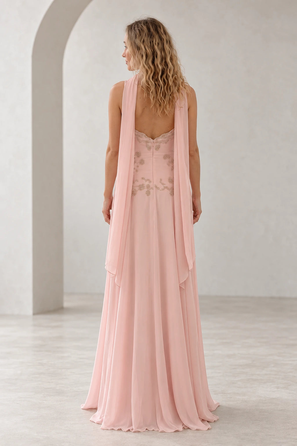 Back view of blush pink chiffon wedding guest dress featuring low lace-trimmed back, floral beaded bodice, flowing scarf panels and long A-line skirt