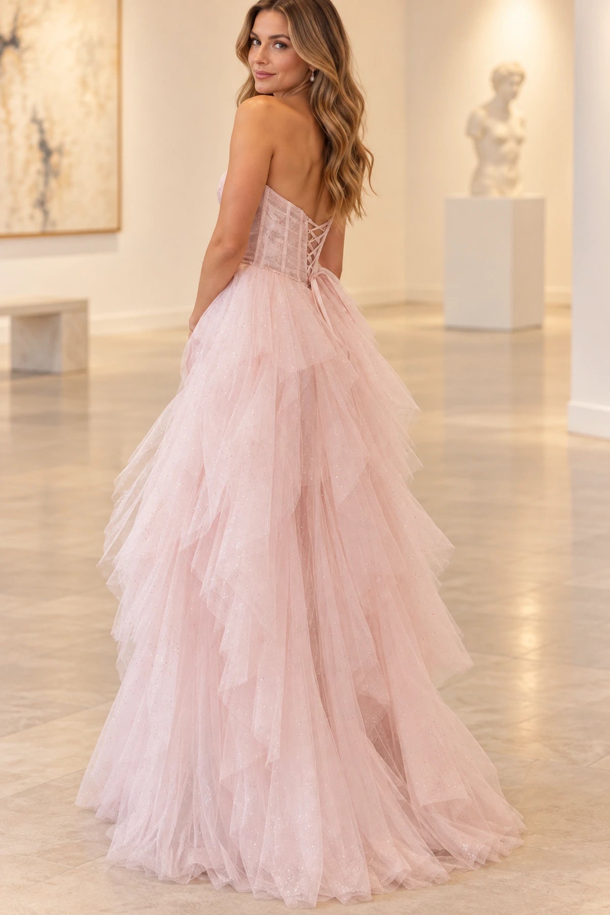 Back view of blush pink strapless princess prom dress featuring a lace-up corset back, delicate waist bow, and flowing layered tulle ruffle skirt.