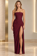 Burgundy strapless sheath prom dress with a straight neckline, asymmetric waist and hip draping, floor-length skirt, and high front slit.