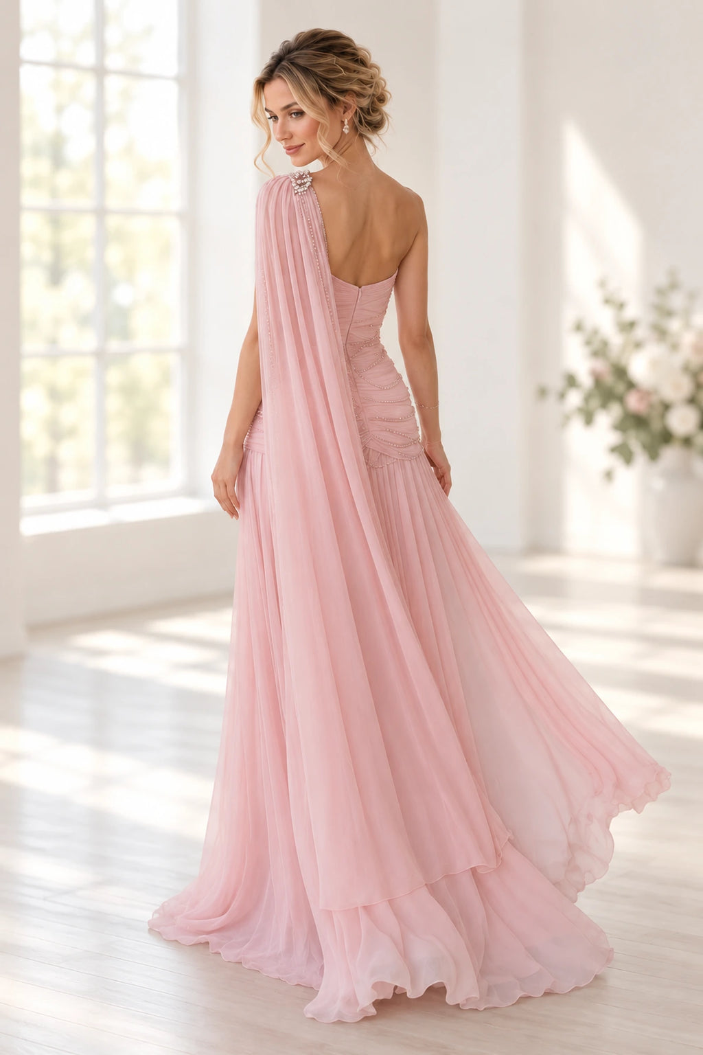 Back view of a blush pink Graceprom wedding guest dress with a low zipper back, ruched beaded bodice and long chiffon cape draped from one shoulder.