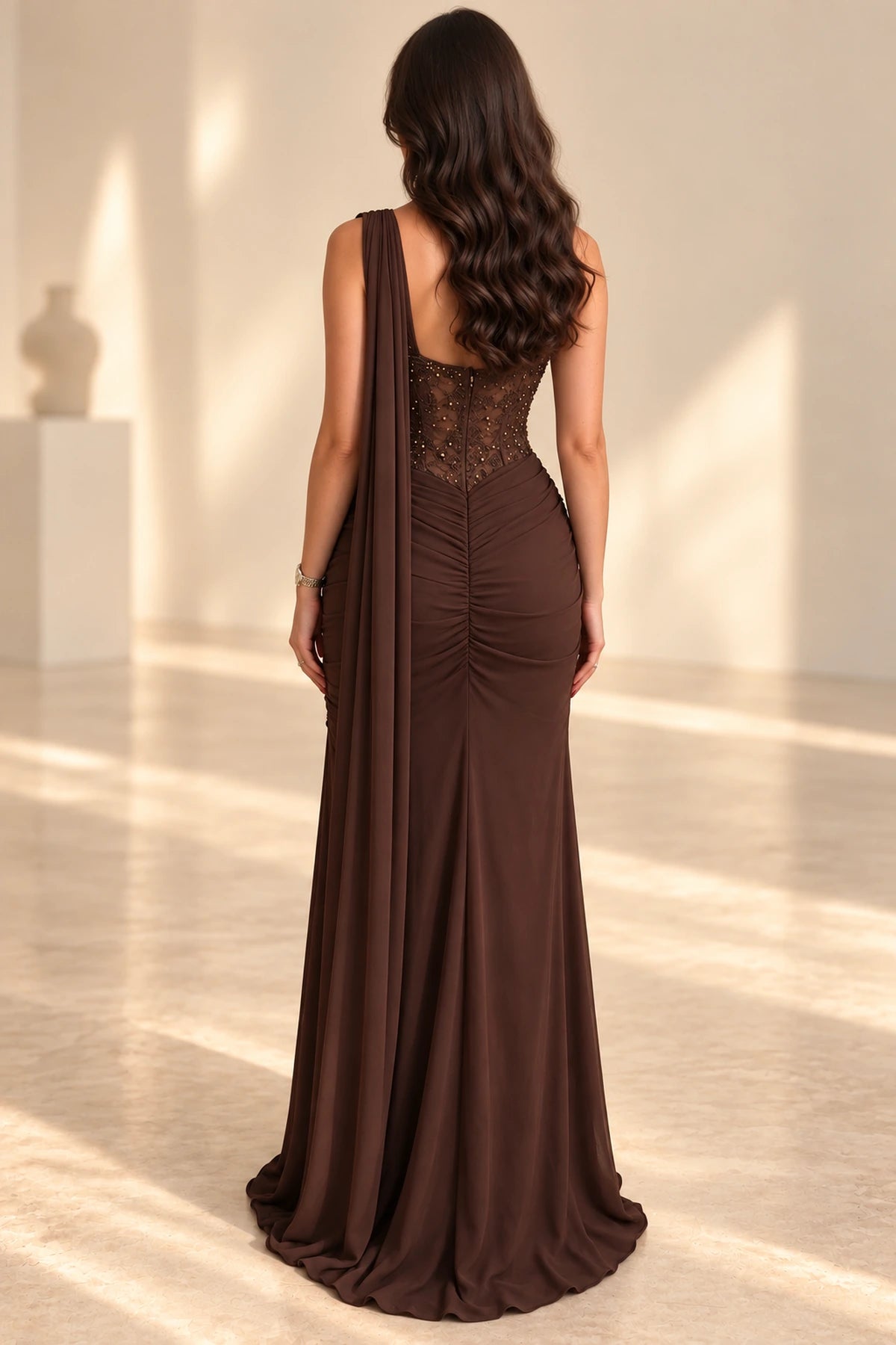 Back view of a chocolate brown chiffon evening gown with a sheer beaded corset back, visible boning, ruched fitted skirt, and long flowing shoulder cape.