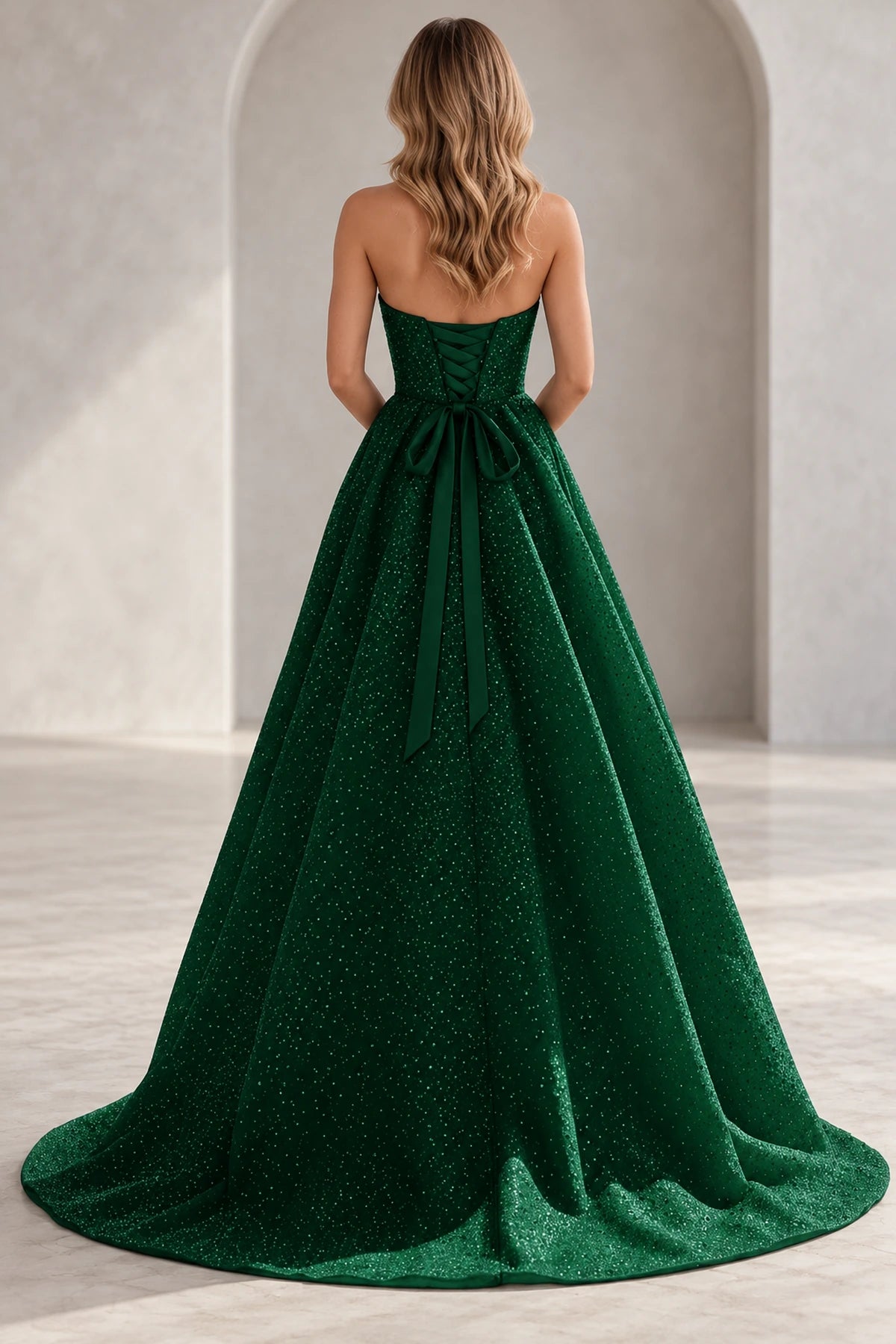 Back view of an emerald green prom dress with corset lacing, a large waist bow, long ribbon tails, and a sparkling skirt with train.