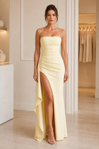 Light yellow strapless floor-length wedding guest dress with ruched bodice, high slit, cascading side drape, and zipper back.
