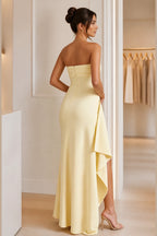 Light yellow strapless floor-length wedding guest dress with ruched bodice, high slit, cascading side drape, and zipper back.