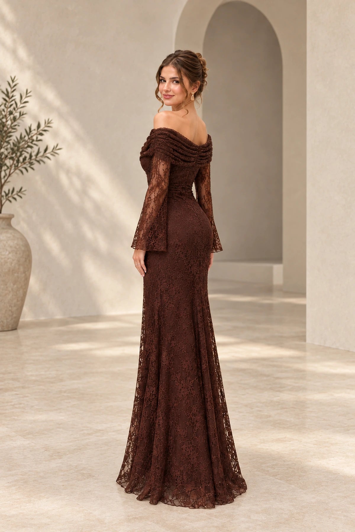 Back view of a chocolate brown lace wedding guest gown with an asymmetric draped neckline, sheer floral lace long sleeves, fitted waist, and flowing floor-length skirt.