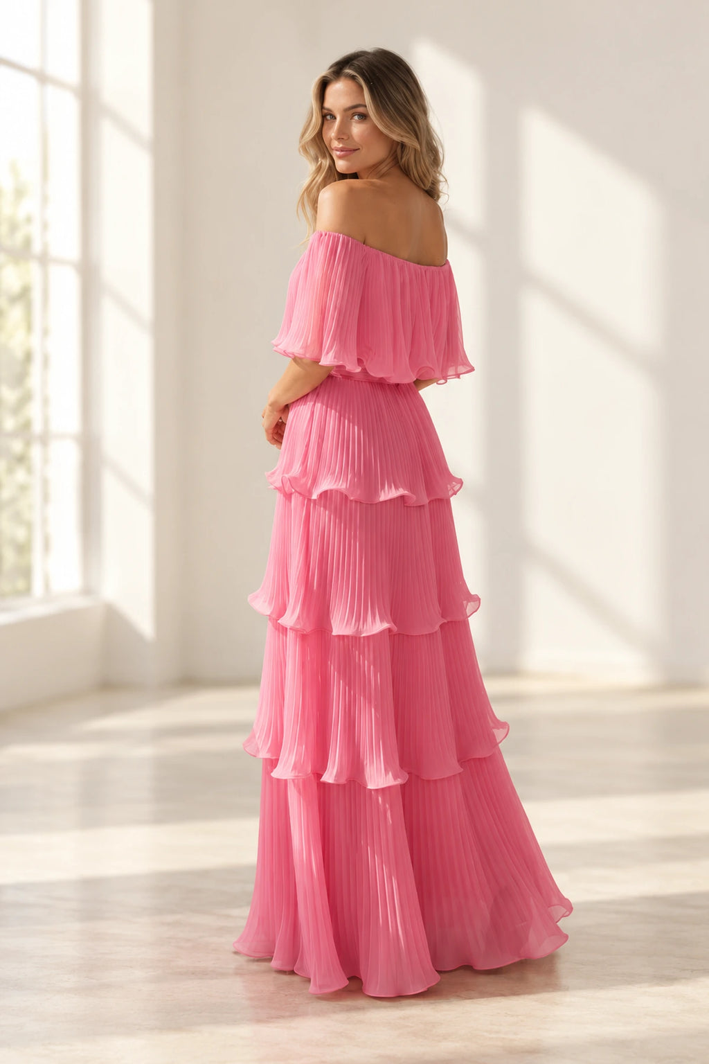 Back view of pink pleated chiffon wedding guest dress featuring off shoulder back, flutter sleeves, fitted waist and layered floor length skirt