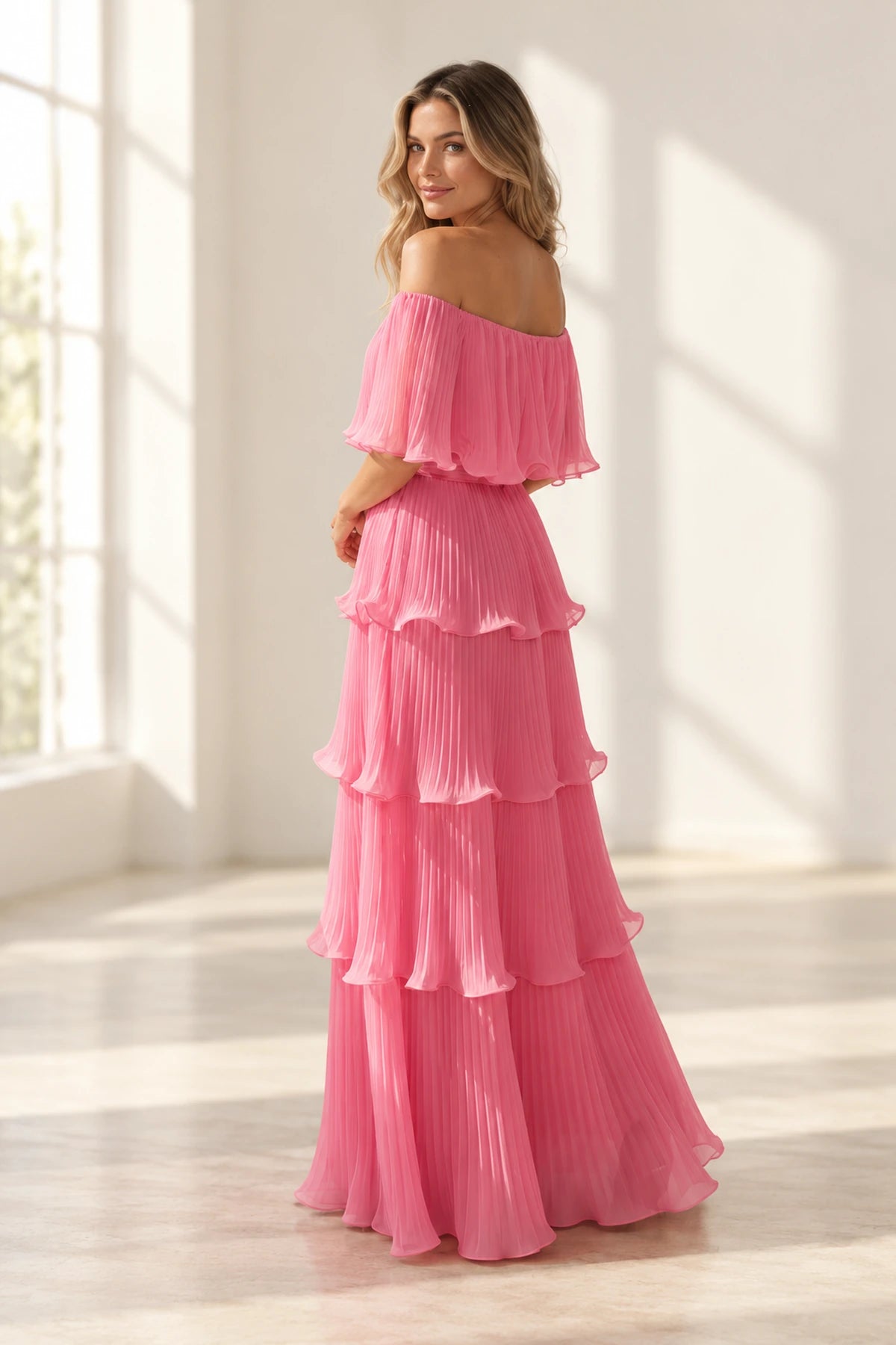 Back view of pink pleated chiffon wedding guest dress featuring off shoulder back, flutter sleeves, fitted waist and layered floor length skirt