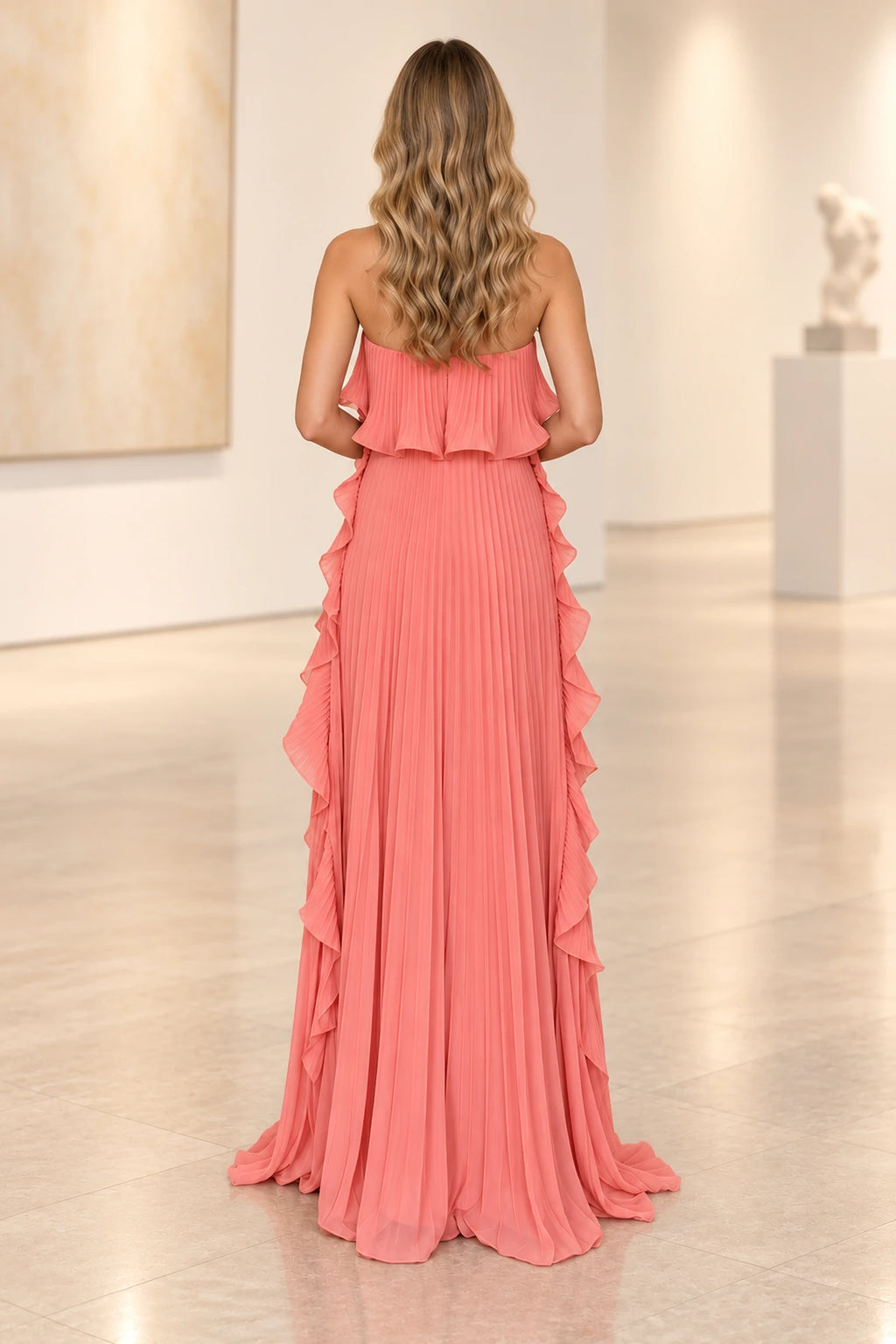 Back view of coral pink pleated chiffon wedding guest dress featuring strapless open shoulder design, ruffled back overlay and long pleated skirt with side ruffles