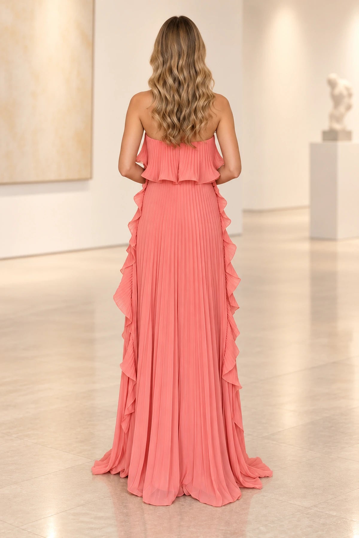 Back view of coral pink pleated chiffon wedding guest dress featuring strapless open shoulder design, ruffled back overlay and long pleated skirt with side ruffles