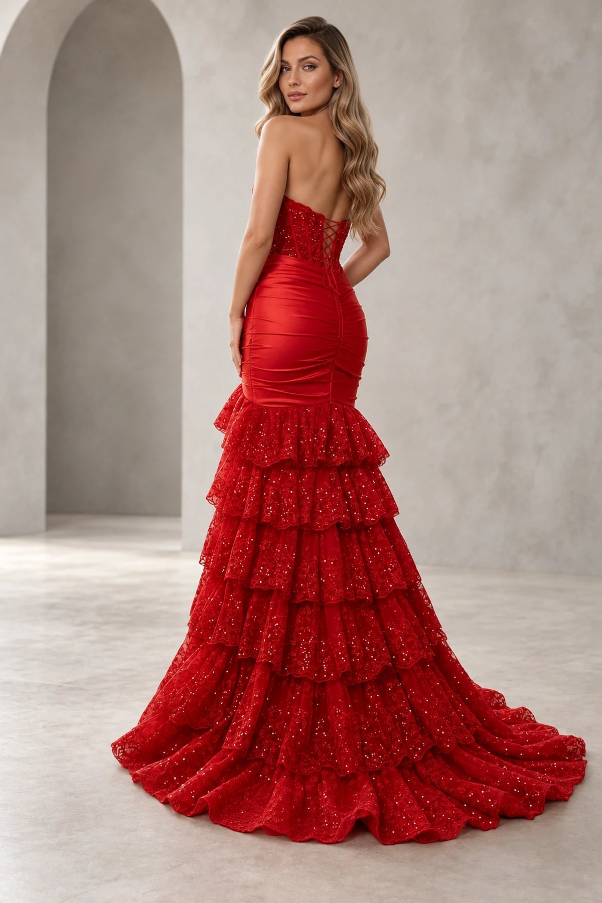 Back view of red mermaid prom dress featuring an adjustable lace-up corset, ruched fitted hips and cascading tiered lace ruffles with a sweeping train.