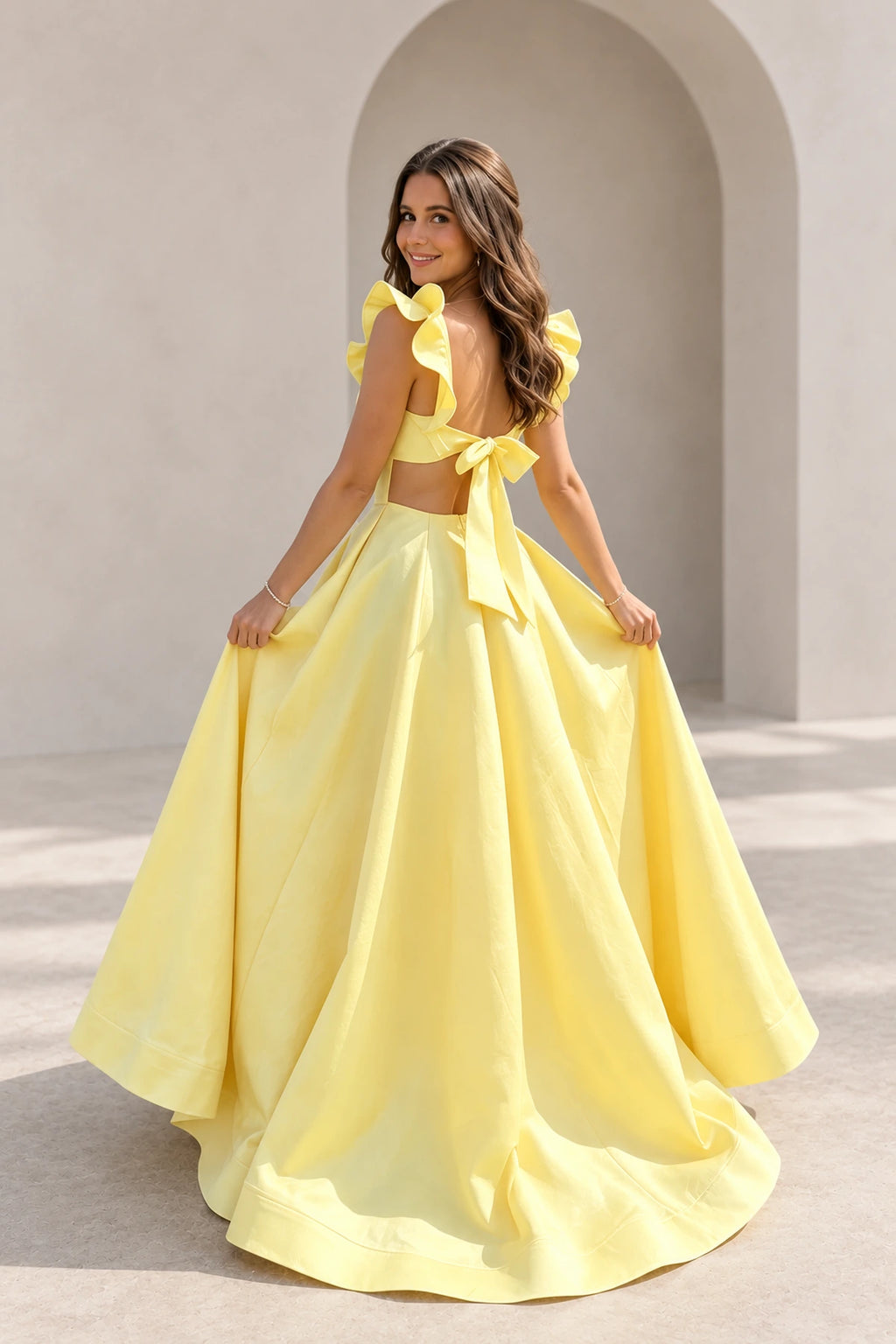 Back view of yellow satin A-line prom dress featuring ruffle shoulder straps, open back with large bow tie and flowing pleated floor length skirt