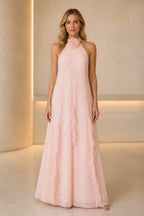 Soft pink A-line wedding guest dress with halter neckline, floral accent, cascading ruffles and floor-length skirt