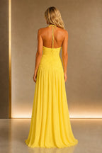 Back view of yellow wedding guest dress with halter tie, open back, ruched bodice and flowing floor-length A-line skirt