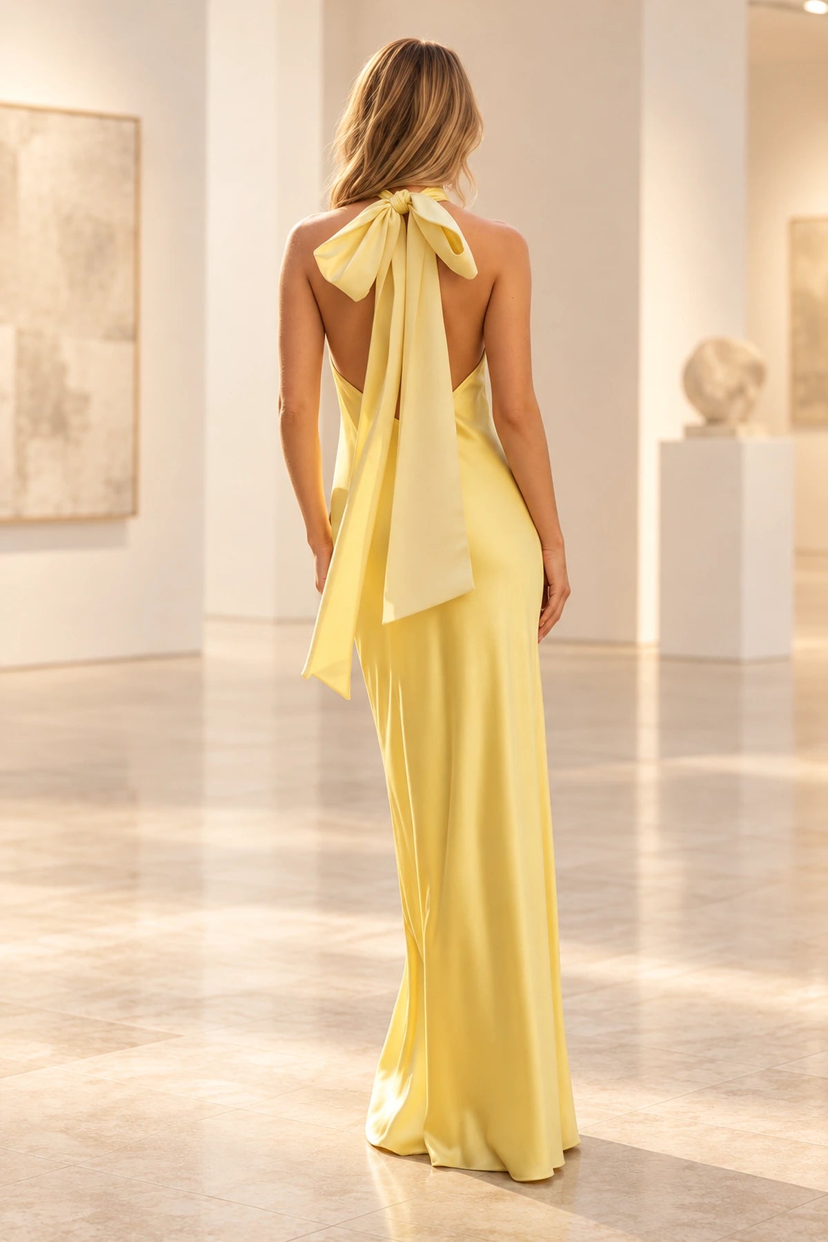 Back view of yellow satin prom dress featuring an open back, oversized halter bow tie, long flowing ribbons, and floor-length sheath skirt.