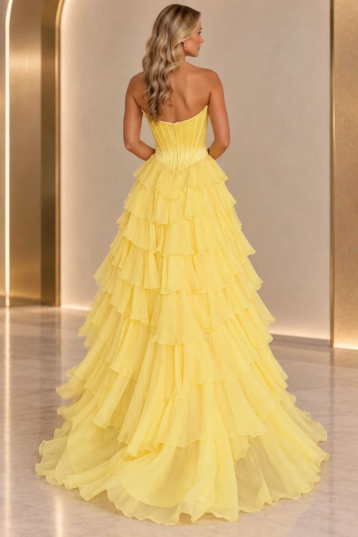 Back view of yellow strapless ball gown prom dress featuring a center zipper, vertically pleated bodice, pointed waistline, and cascading floor-length ruffles.